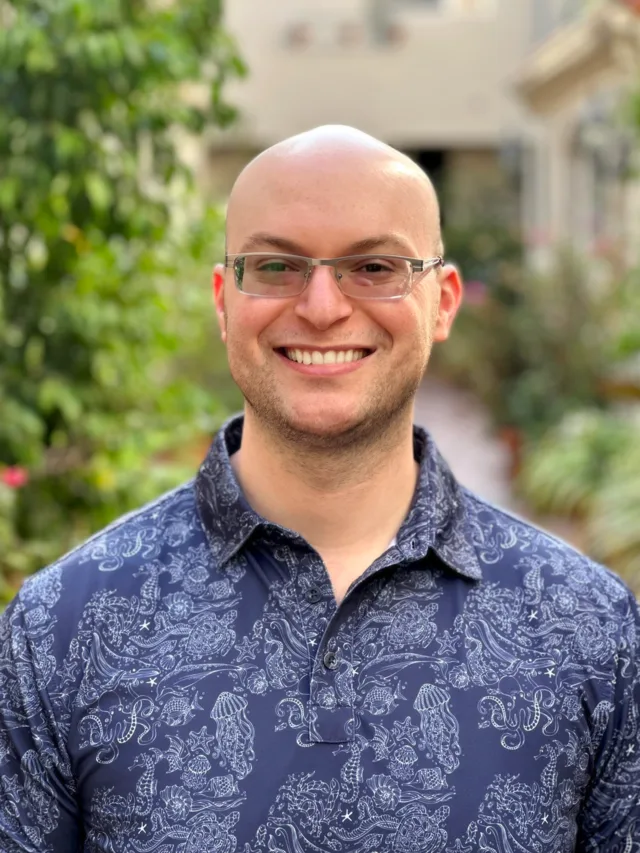 Ben Reicher, smiling, wearing glasses and a blue shirt with a white marine life pattern, outdoors